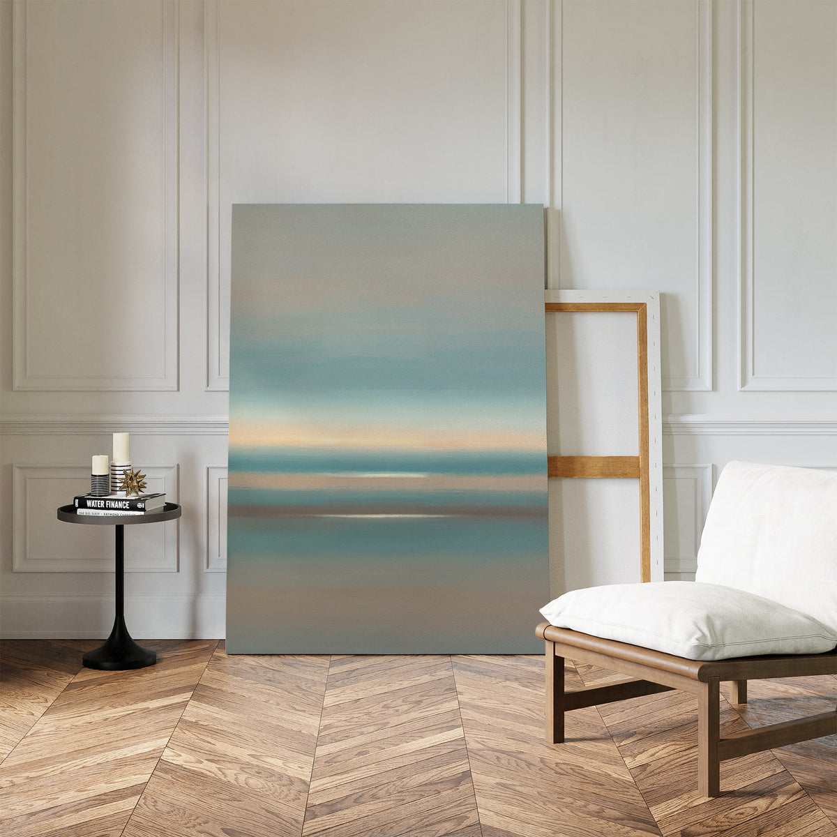 A minimalist room featuring chevron wood floors, a white cushioned chair, a black round table with candles and books, and the Soft Tide Layers Canvas Art - Vertical leaning against a white paneled wall for tranquil home decor.
