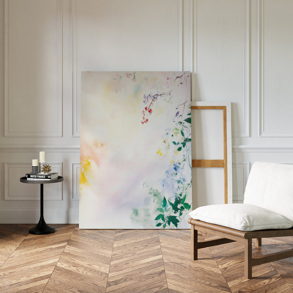 The "Spring Reverie Canvas Art - Vertical," featuring soft pastel florals, leans against a white paneled wall beside an empty frame, with a cushioned chair and round side table on a herringbone wood floor.