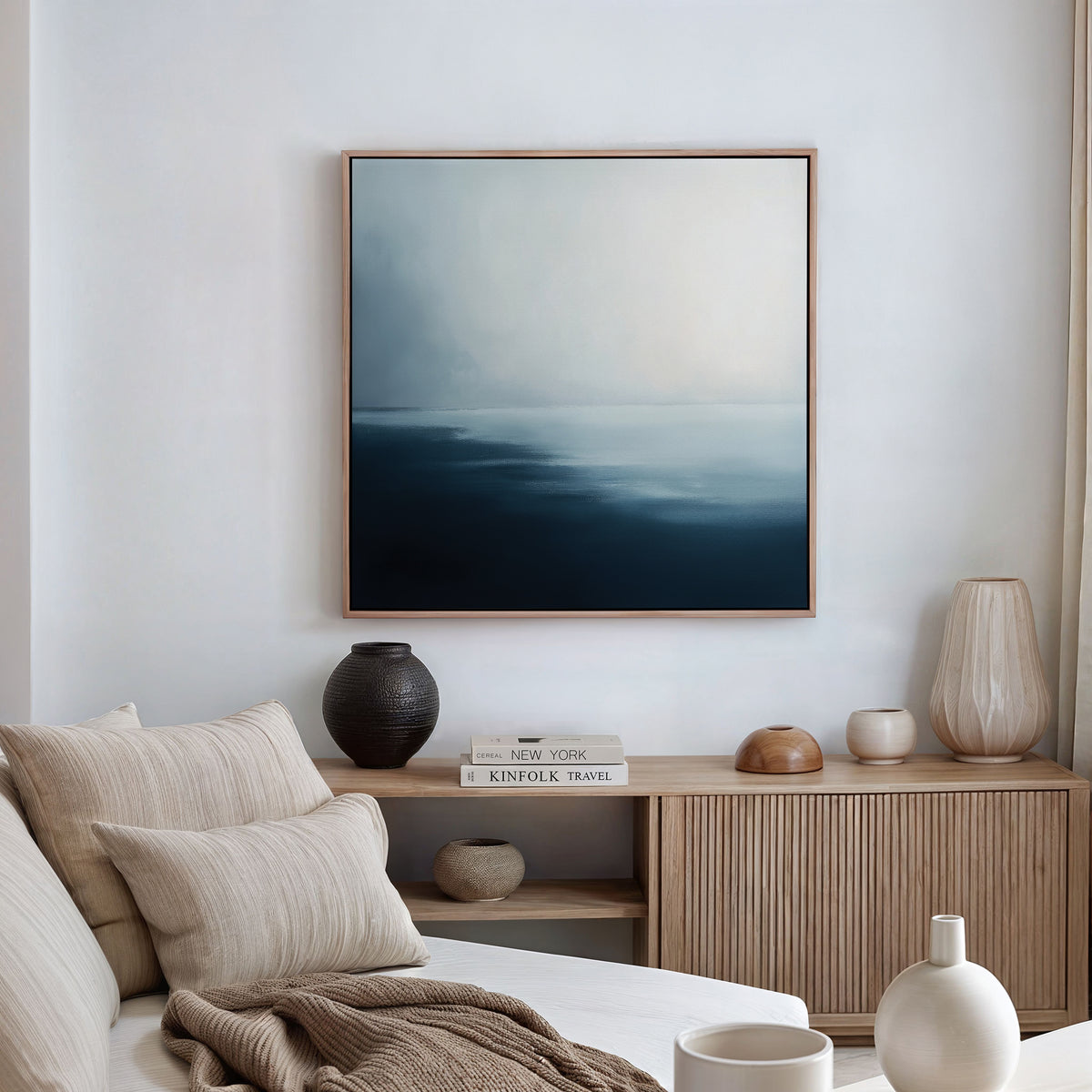 A minimalist living room with a beige sofa, neutral pillows, a wooden sideboard decorated with accents, and the Steel Harbor Mist Canvas Art - Square in white and gray tones displayed above the sideboard.