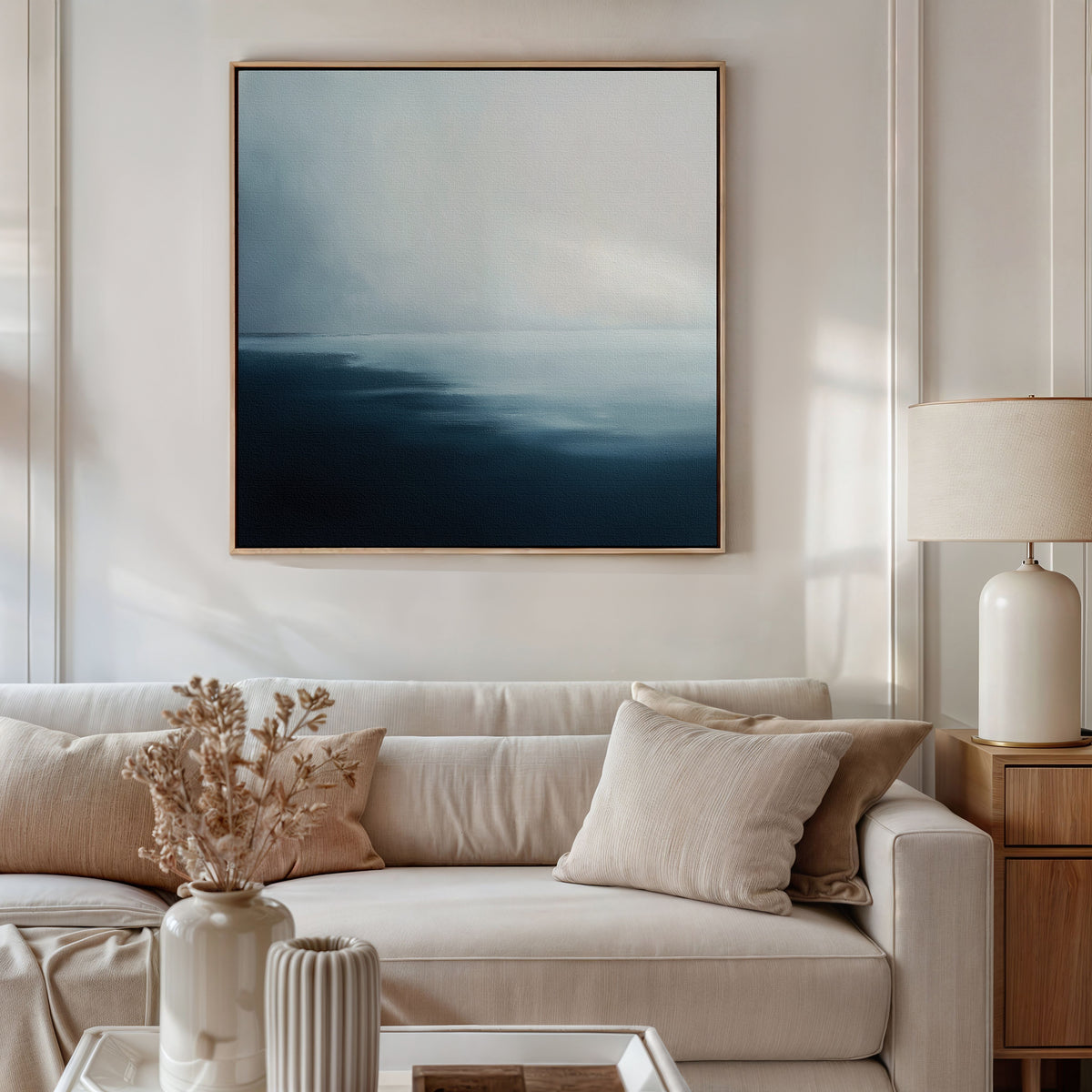 A modern living room with a beige sofa, neutral pillows, a ceramic vase with dried flowers, a table lamp, and the Steel Harbor Mist Canvas Art - Square—a striking abstract blue-and-white painting—hanging above the sofa.