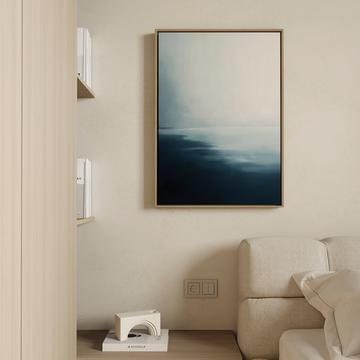 A minimal living room with a light sofa, wooden shelf of white books, and the Steel Harbor Mist Canvas Art - Vertical—an abstract blue and white framed piece. A small geometric decor accent rests on the floor.