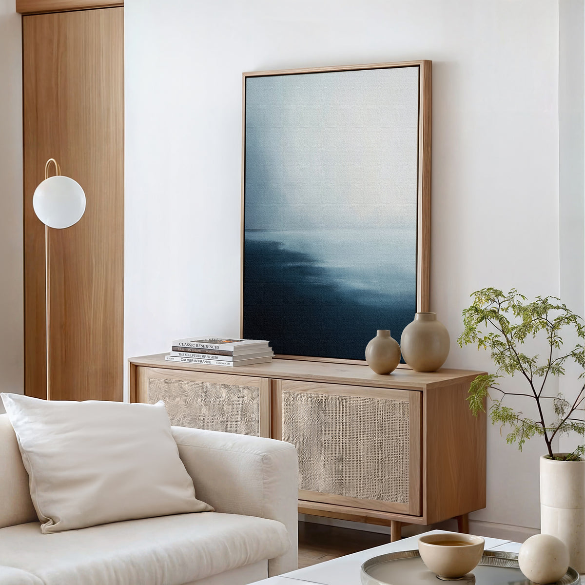 A minimalist living room features the Steel Harbor Mist Canvas Art - Vertical, neutral pottery, stacked books, a light wood sideboard, a white sofa with a cushion, a round lamp, and a small green plant.