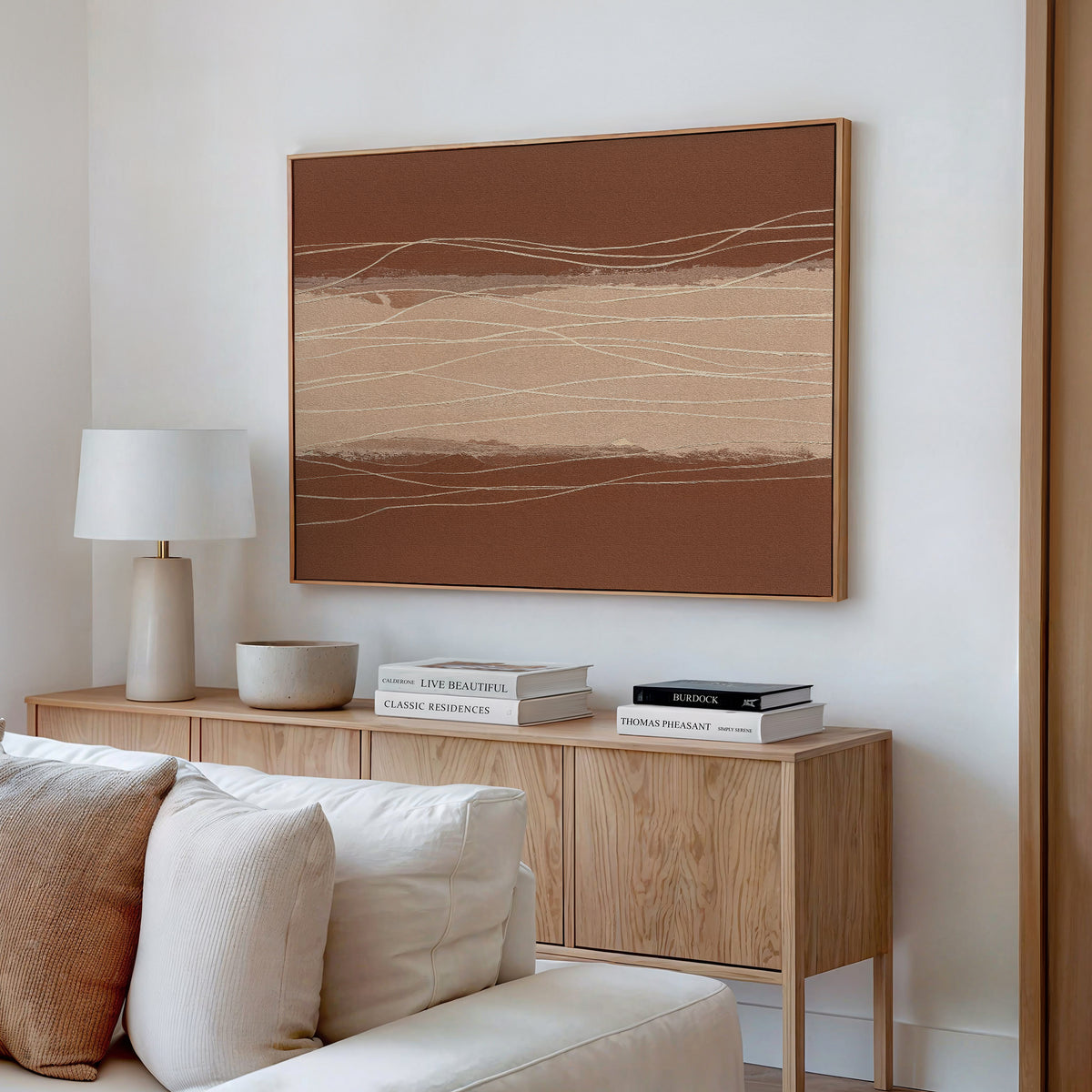 A modern living room features a wooden sideboard with stacked books, a ceramic bowl, a white lamp, and the Terracotta Currents Canvas Art - Horizontal displayed above a light-colored sofa.