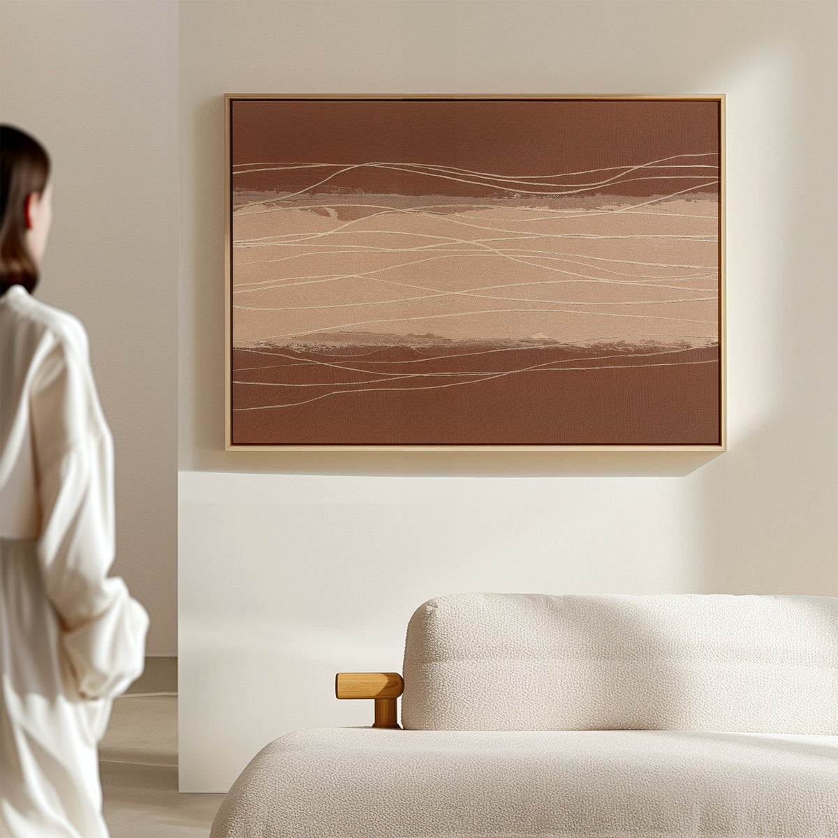 A person in white stands in a minimal, bright room, gazing at the Terracotta Currents Canvas Art - Horizontal with abstract brown and beige lines, displayed above a textured, light-colored sofa.