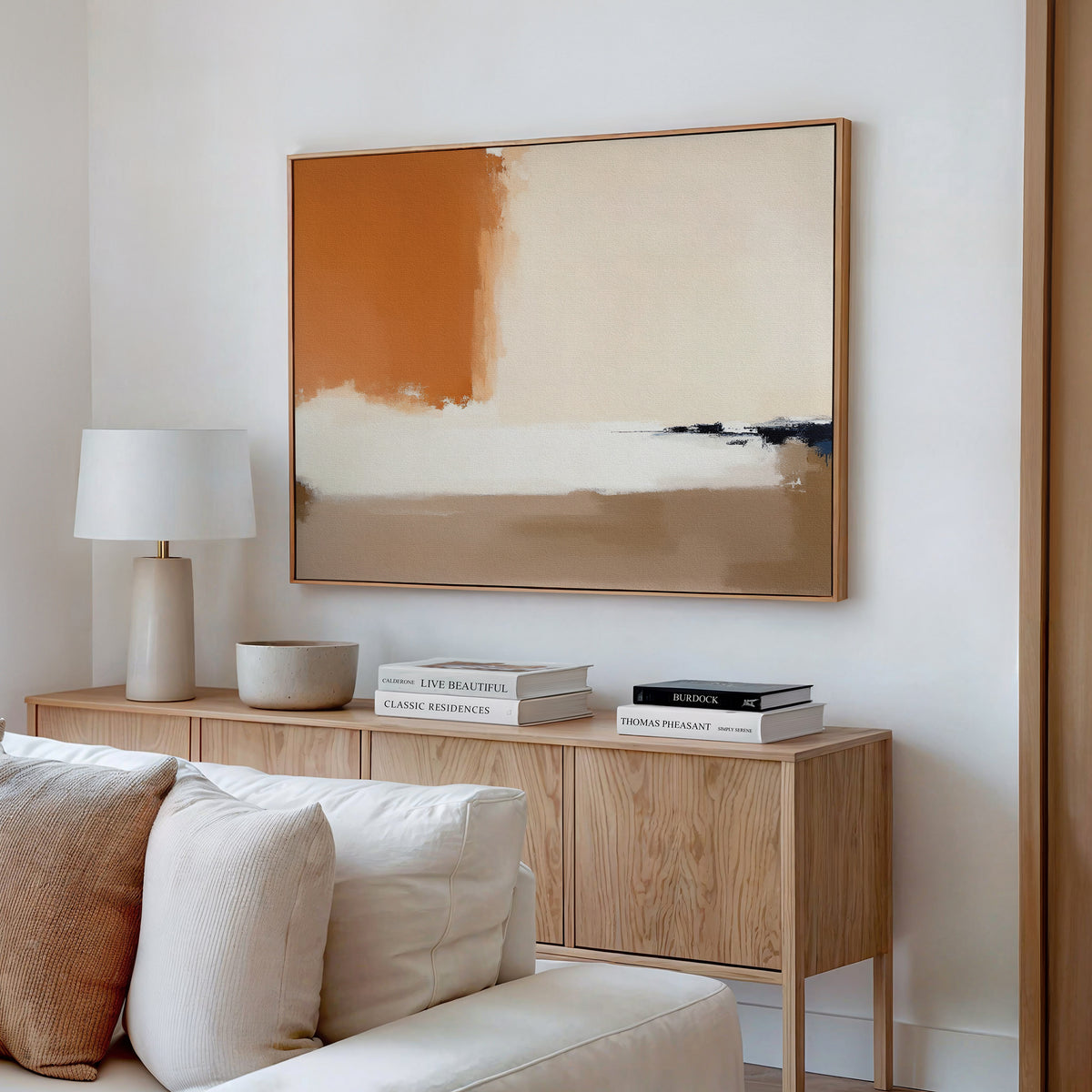 A modern living room featuring the Terracotta Edge Canvas Art - Horizontal above a wooden sideboard with neutral decor, a white lamp, stacked books, and a cream-colored sofa partially visible in front.