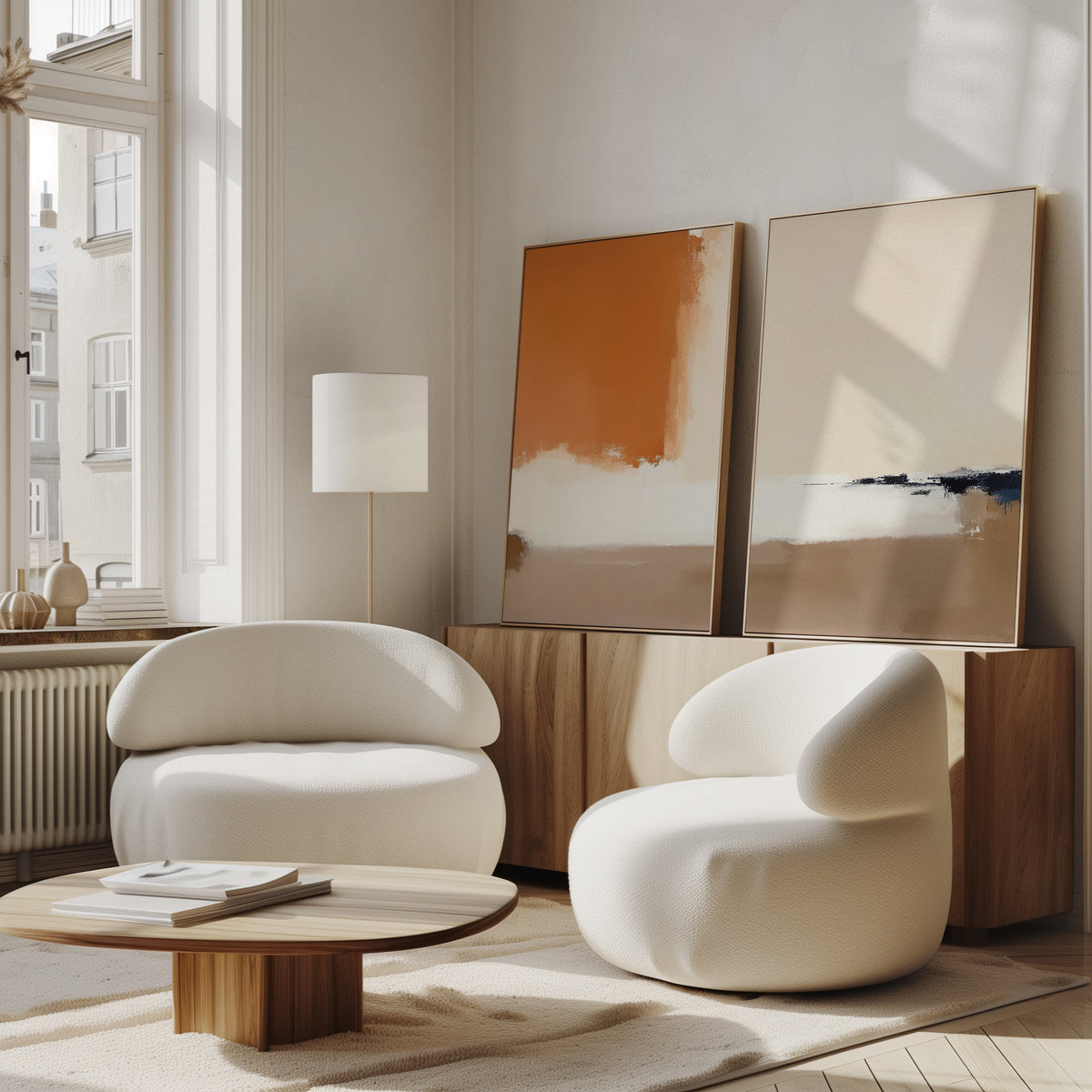 A modern living room features the Terracotta Edge - Set Of 2 white, rounded armchairs, a low wooden coffee table, and an abstract print by a wooden sideboard. Sunlight streams through large windows, highlighting the stylish decor.