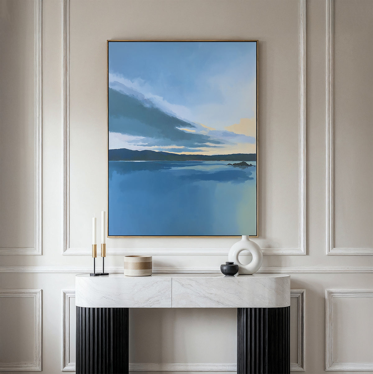 The Tranquil Reflections Canvas Art - Vertical, featuring a serene blue seascape, hangs on a beige wall above a white marble console table with black legs, adorned with minimalist vases and candle holders.