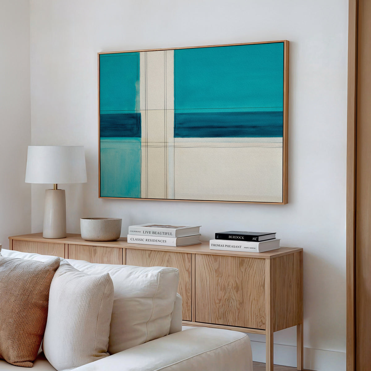 A modern living room features a light wood sideboard, stacked books, a white lamp, a bowl, and the Turquoise Grid Canvas Art - Horizontal above a cream sofa with neutral cushions for striking modern decor.
