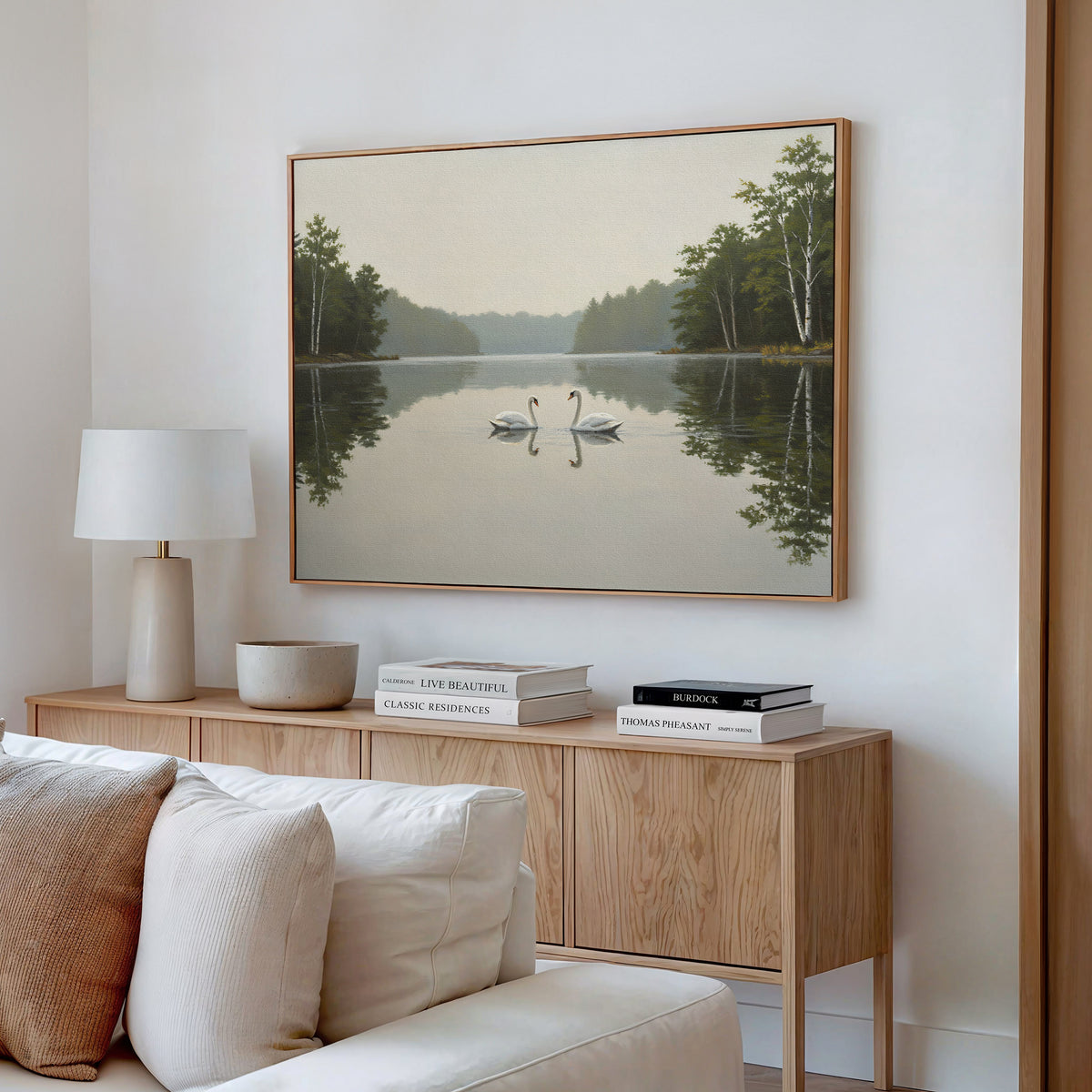 A serene living room features a light wood sideboard, white lamp, and books. Above, the Twin Swans Serenity Canvas Art - Horizontal depicts two swans on a calm lake, adding a nature-inspired touch to the decor.