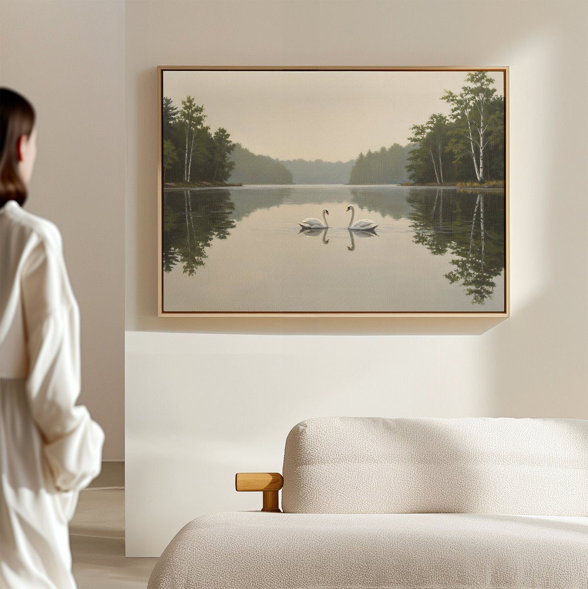 In a minimal bedroom, a person gazes at the Twin Swans Serenity Canvas Art—featuring two swans on a calm lake with trees—framed and hanging above a light-colored bed.