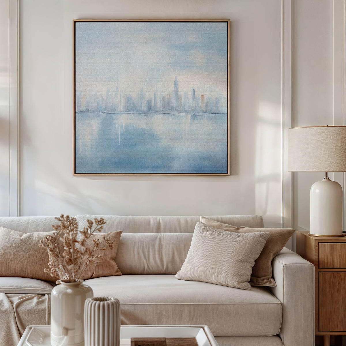 A modern living room with a light beige sofa, neutral pillows, a side table topped with a cream lamp, and vases of dried flowers. Above the sofa hangs the Urban Mirage Canvas Art - Square, adding striking modern cityscape wall decor.
