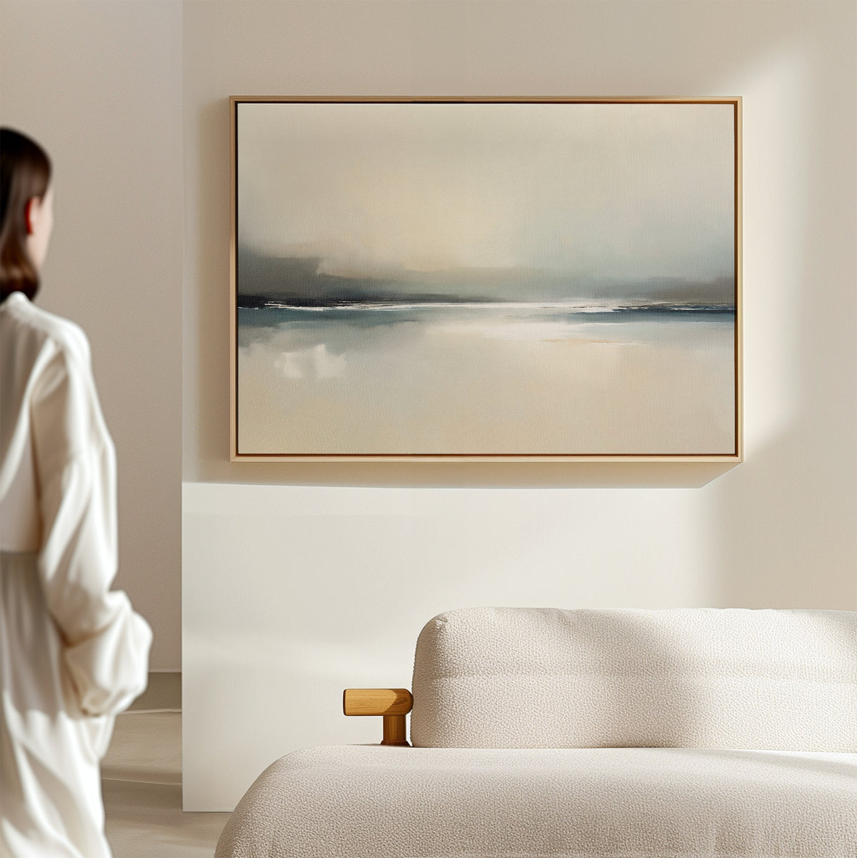 A person in a white shirt stands in a minimalist room, gazing at the Veiled Estuary Canvas Art - Horizontal with soft blue and beige tones displayed above a beige sofa, creating a calm and serene atmosphere.