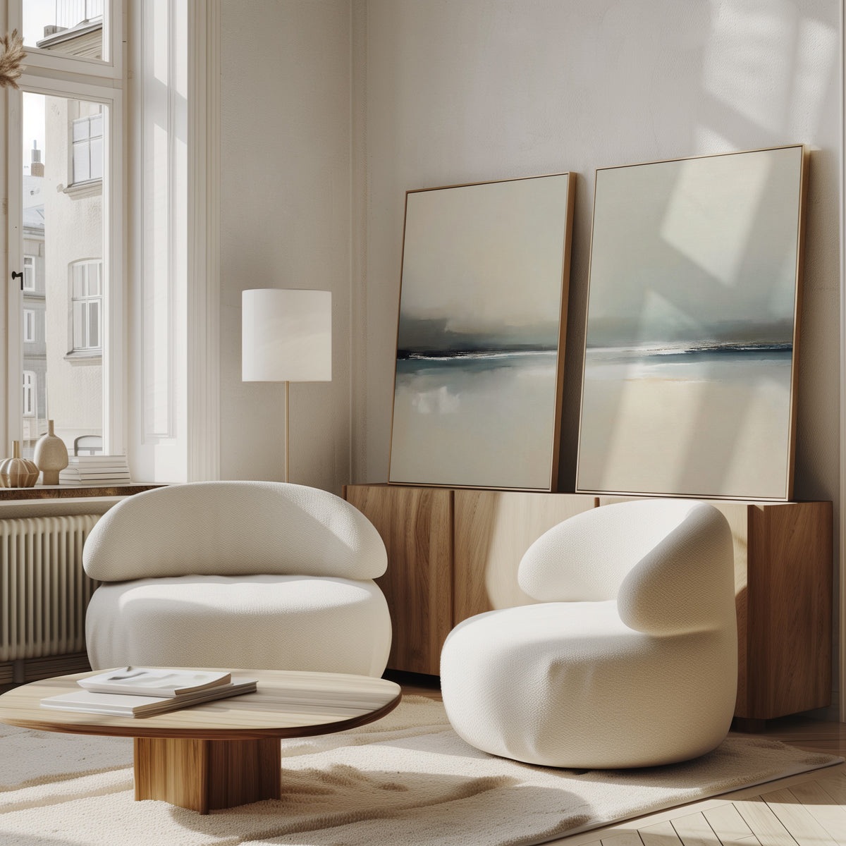 A modern living room with two rounded white armchairs, a low wooden coffee table, a wooden sideboard, and the "Veiled Estuary - Set Of 2" contemporary wall art leaning against the wall, illuminated by large windows letting in natural light.
