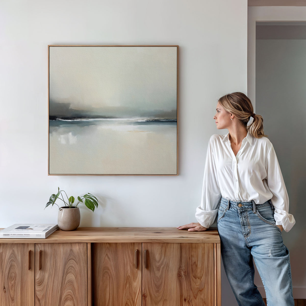 A woman in a white blouse and jeans leans on a wooden cabinet, gazing at the Veiled Estuary Canvas Art - Square on the wall. A plant and books sit on the cabinet beneath this serene, contemporary artwork.