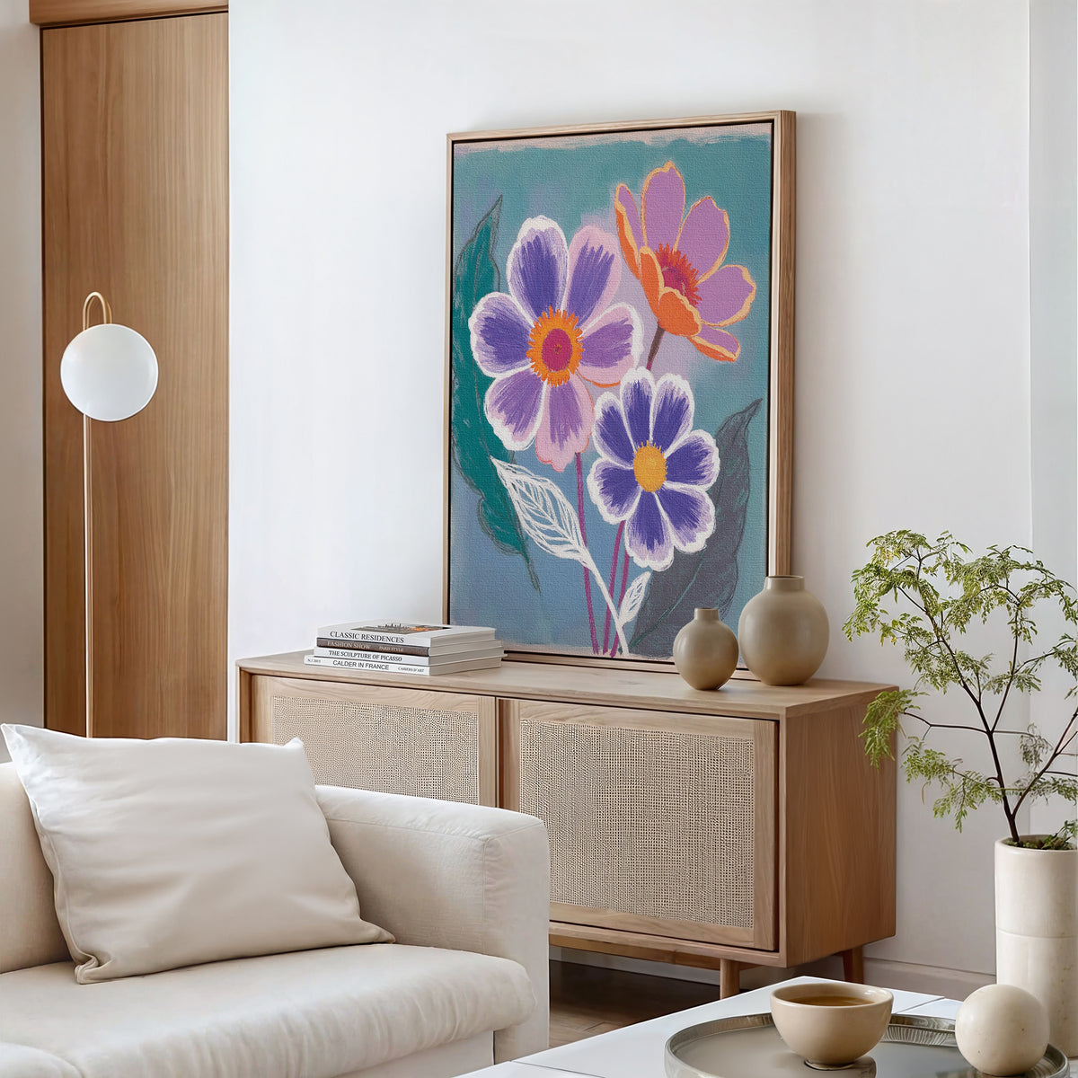 A modern living room with a light wood sideboard, Vibrant Petals Canvas Art - Vertical, neutral vases, books, a small green plant, and a white cushioned chair with a pillow in the foreground.