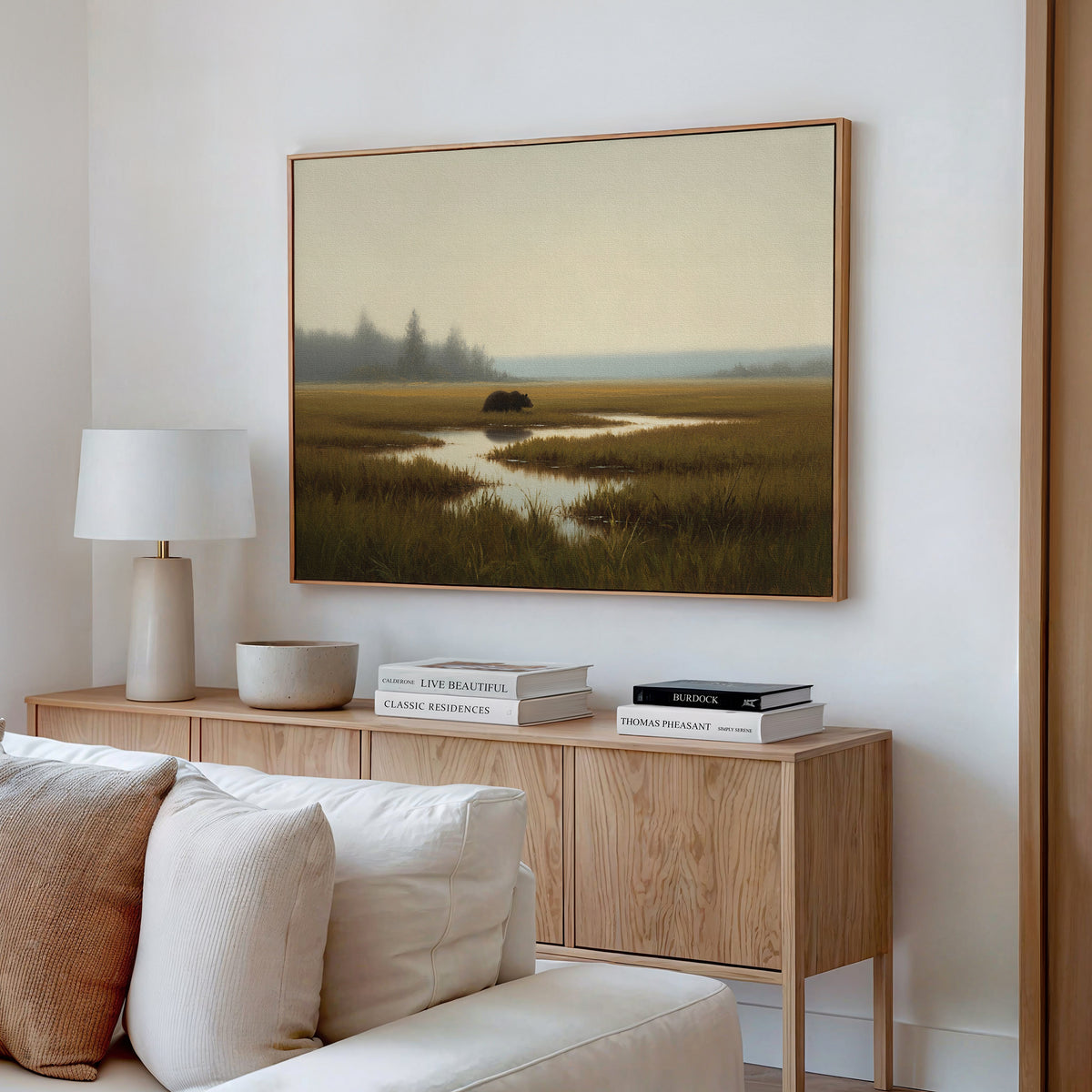 A modern living room with a light wood console, white lamp, neutral decor, and a white sofa. Above the console hangs the Wetland Bear Canvas Art - Horizontal featuring a tranquil misty marsh with distant trees and calm water.