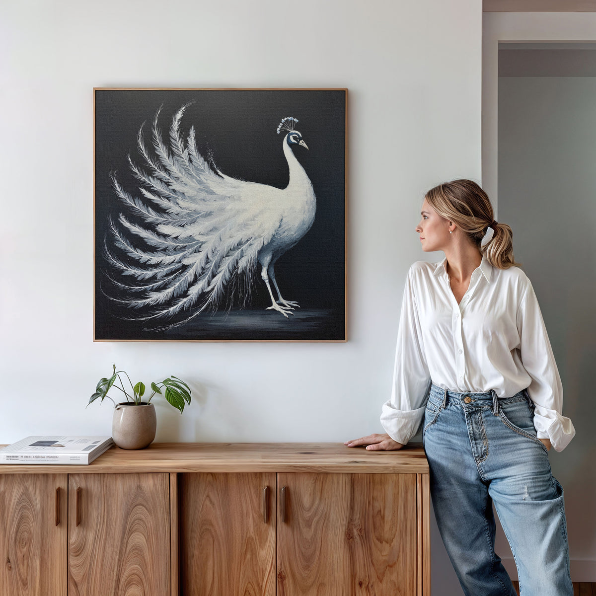 A woman in a white shirt and jeans leans against a wooden cabinet, gazing at the White Peacock Display Canvas Art - Square that hangs on the white wall. A potted plant and books add decor to the cabinet.