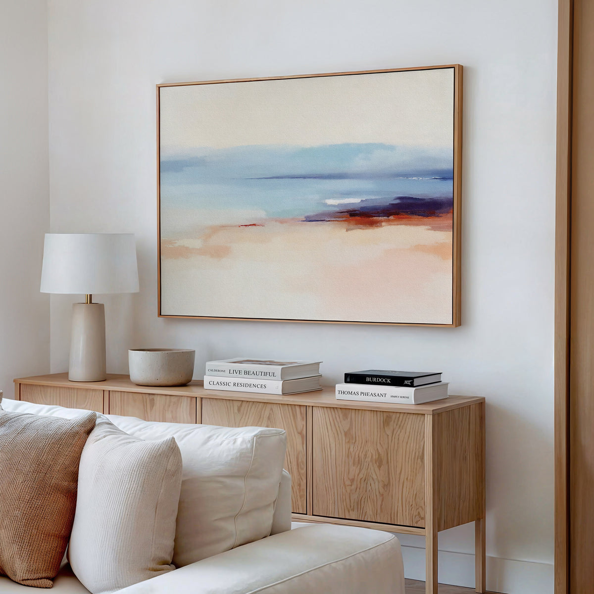 A minimalist living room with a wooden sideboard, neutral decor, stacked books, a white lamp, and the Windswept Coastline Canvas Art - Horizontal featuring soft pastel hues displayed on a white wall.