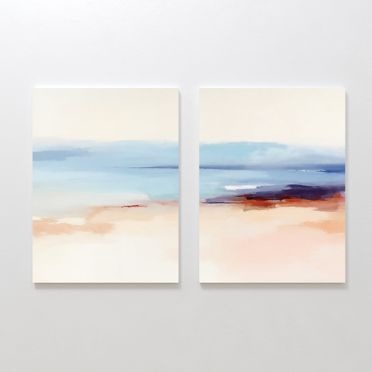 The "Windswept Coastline - Set Of 2" features abstract paintings in blue, beige, and peach tones, evoking a serene seascape. Hung side by side, these prints create elegant coastal wall art on a white wall.