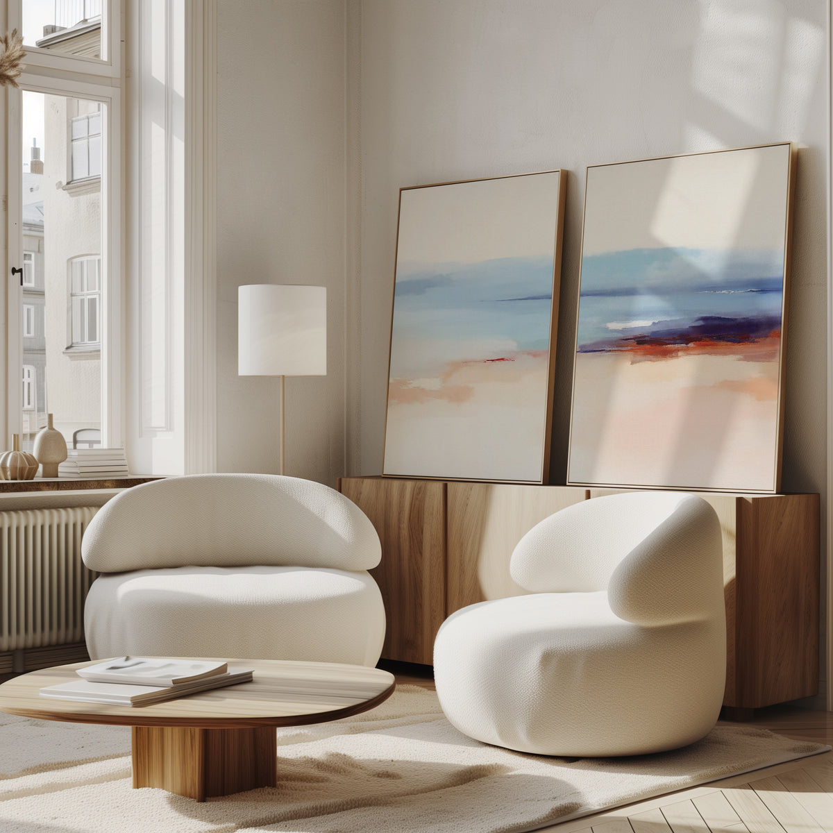 A modern living room features two rounded white armchairs, a low wooden coffee table, a floor lamp, and the Windswept Coastline - Set Of 2 wall art set atop a wooden sideboard against a white wall, brightened by large windows.