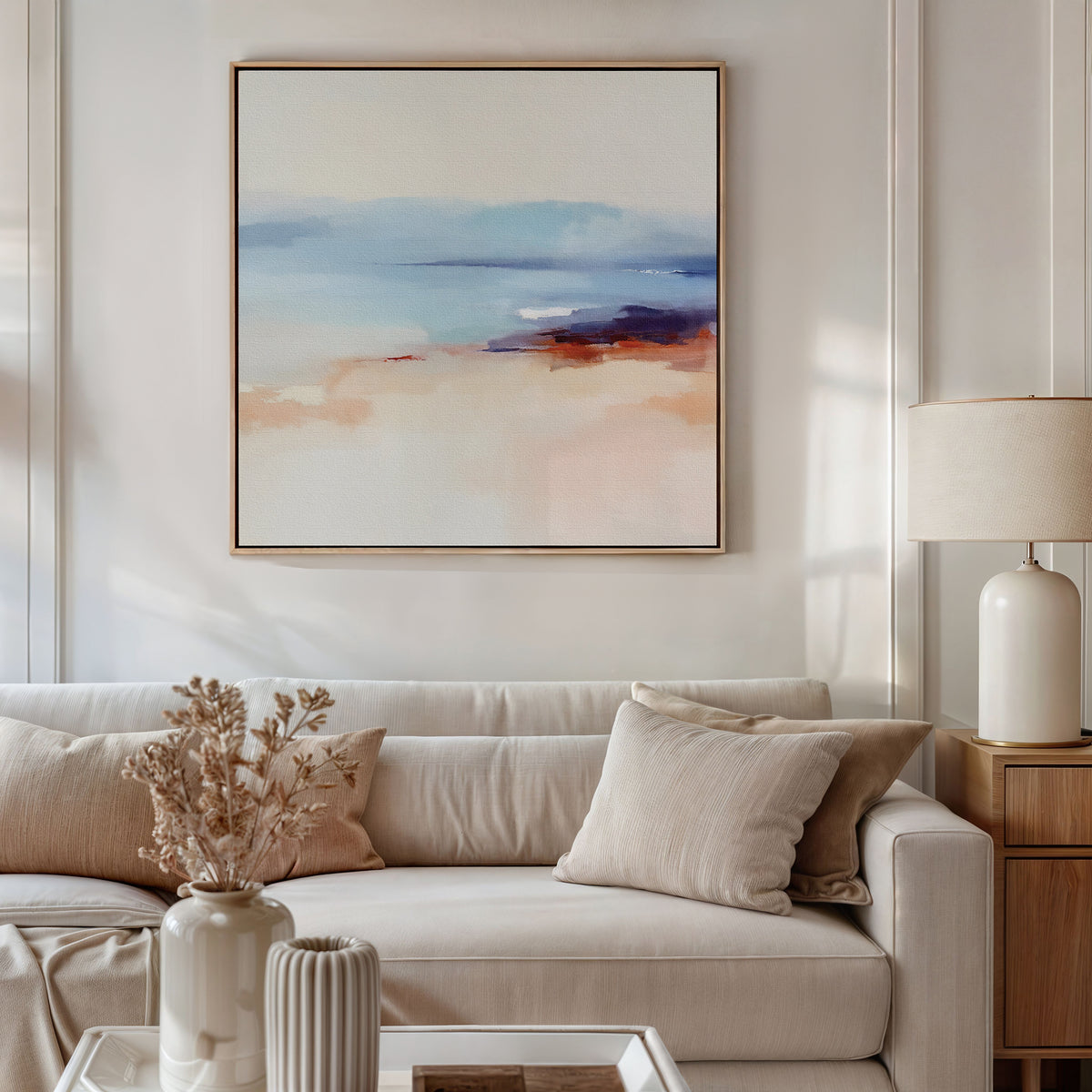 A modern living room with a beige sofa, light cushions, dried flowers in a vase, and a table lamp on a wooden side table, featuring the Windswept Coastline Canvas Art - Square in blue, peach, and cream tones for a tranquil ambiance.