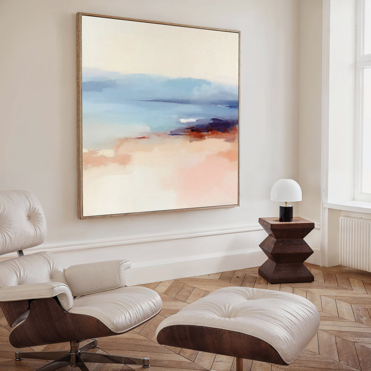 A modern living room featuring a white leather lounge chair with ottoman, a wooden side table with dome lamp, and the Windswept Coastline Canvas Art - Square in soft blue, peach, and cream tones displayed on the wall.