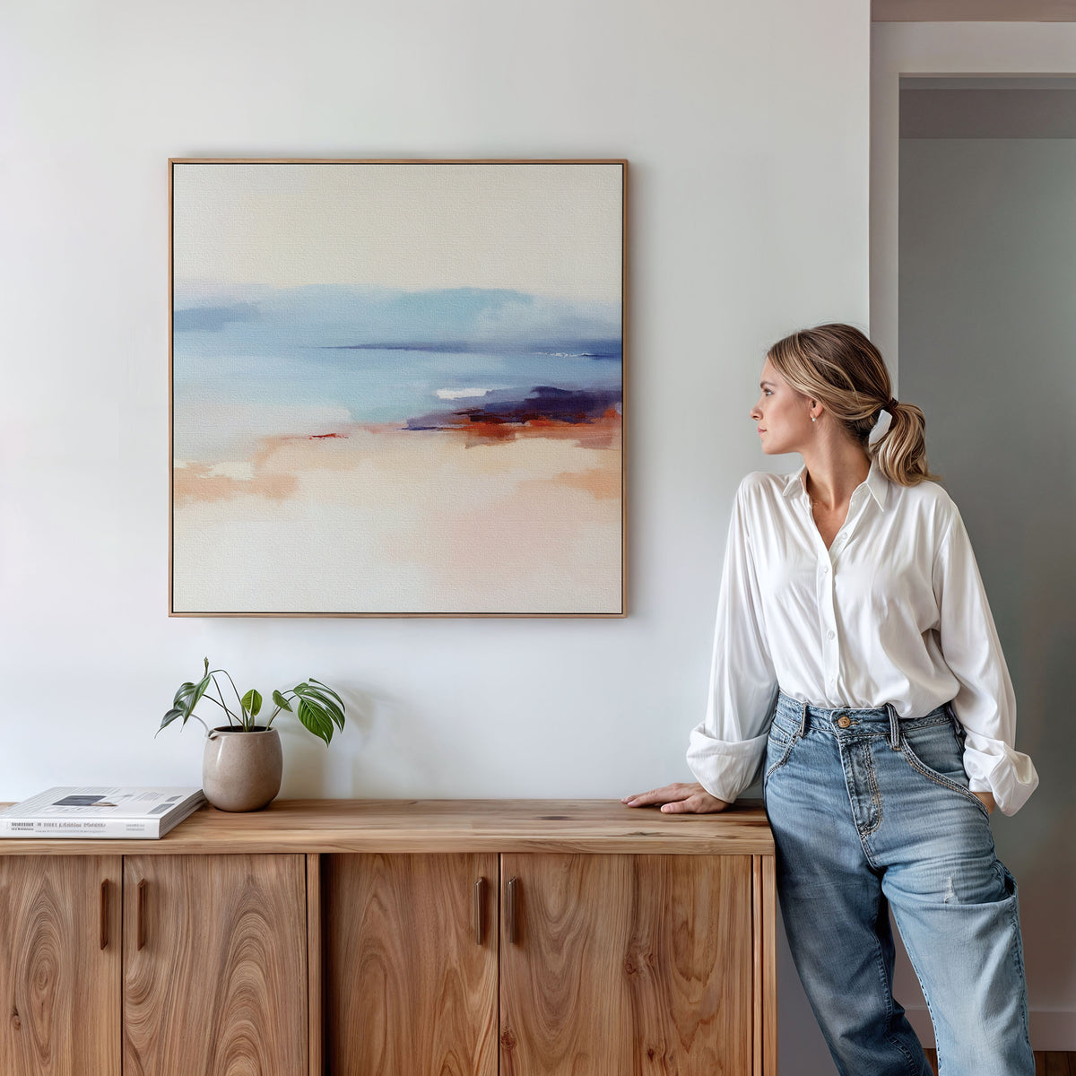 A woman in a white blouse and jeans leans against a wooden cabinet, gazing at the Windswept Coastline Canvas Art - Square with soft pastel colors. A potted plant and book rest on the cabinet below the serene coastal print.