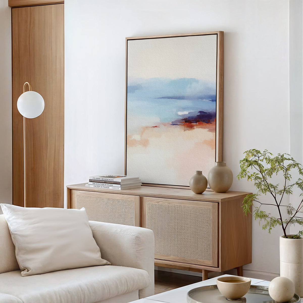 A modern living room with a light wood cabinet, the Windswept Coastline Canvas Art - Vertical as wall art, ceramic vases, books, a small plant, a round hanging lamp, and a white sofa with a cream pillow.
