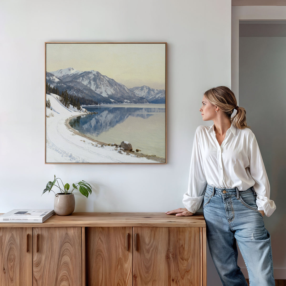 A woman in a white shirt and blue jeans stands in a modern room, leaning on a wooden sideboard and admiring the Winter Reflection Canvas Art - Square, which features mountains, snowy shores, and a calm alpine lake—a perfect winter scene.