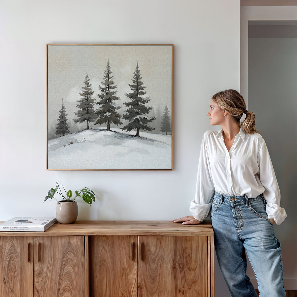 A woman in a white blouse and jeans stands by a wooden console with a small plant and books, admiring the Winter Solitude Canvas Art - Square featuring pine trees on a white wall.