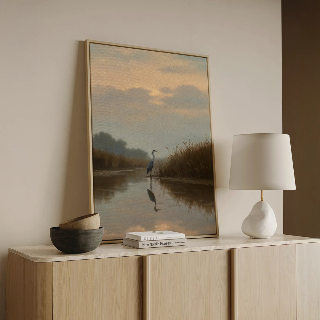 A framed painting of a heron by a calm river rests on a light wood cabinet beside a modern white lamp, a small bowl, and stacked books in a minimalist room.