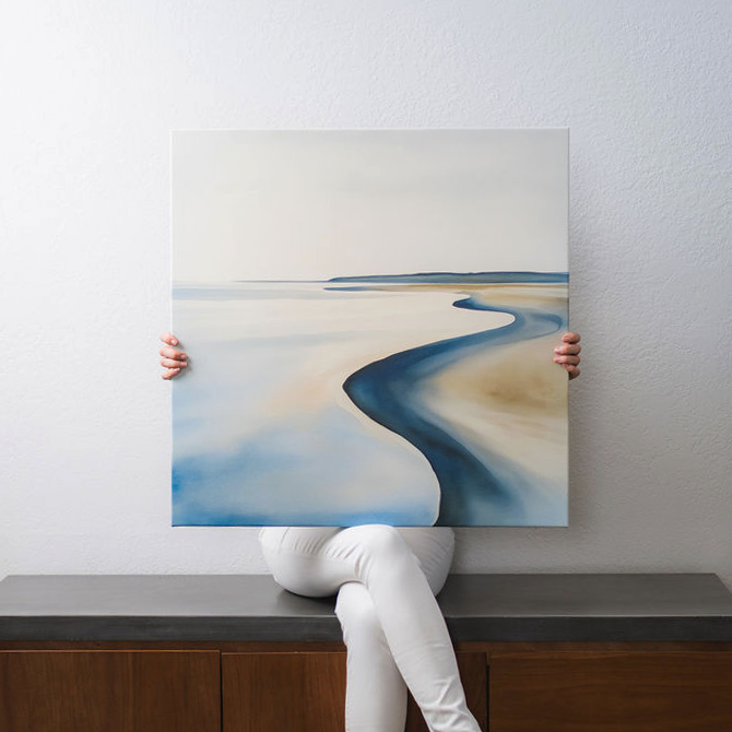A person dressed in white sits on a bench, holding a square painting in front of their face. The painting depicts a minimalist seascape with a winding shoreline.
