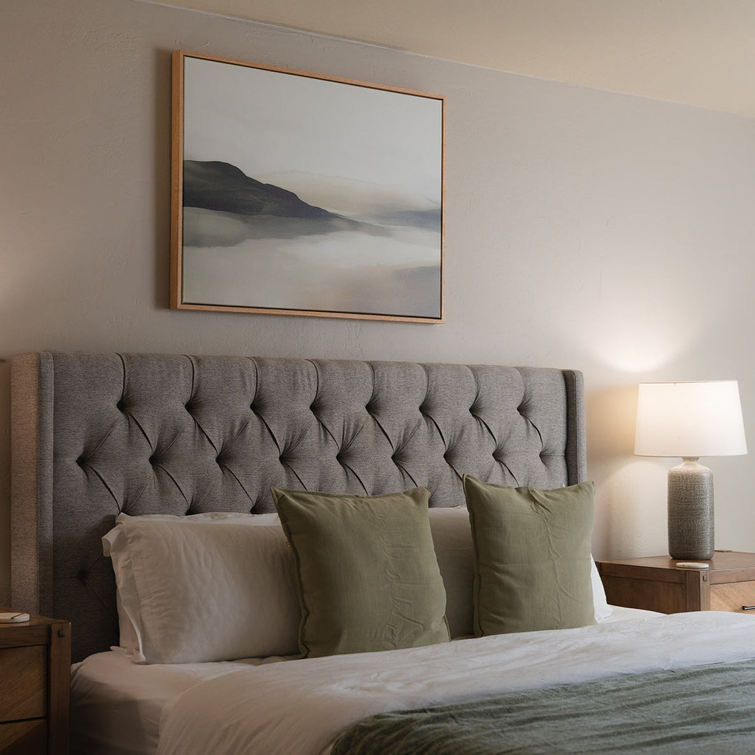 A minimalist bedroom with a beige bed, white pillows, and a soft, neutral wall. Above the bed hangs a framed abstract landscape painting in muted tones. A bare branch decorates the nightstand.
