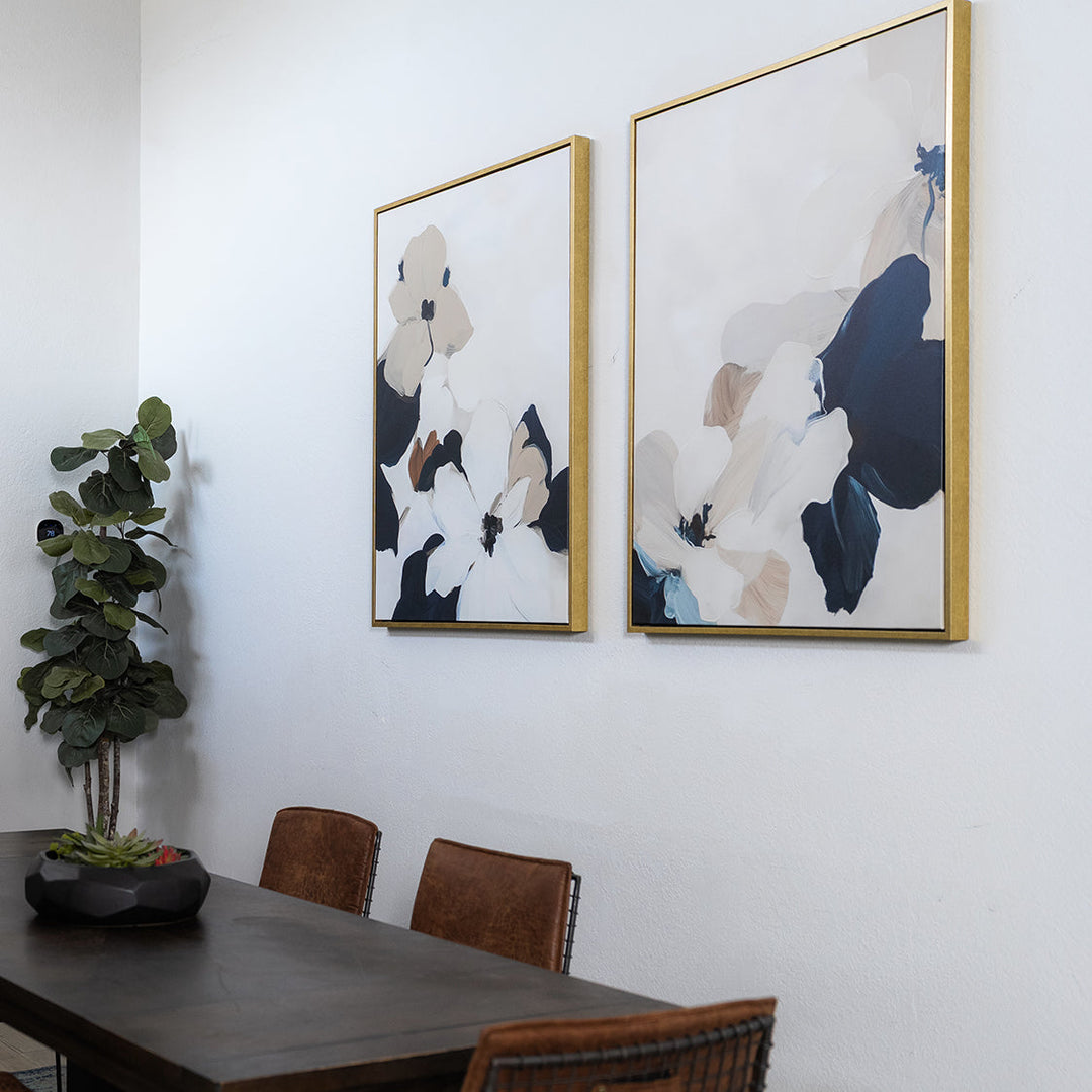 A modern dining area with a dark wooden table, brown chairs, a potted plant, and two abstract floral paintings in gold frames hanging on a white wall.