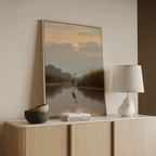 The Misty Reflection Canvas Art features a heron in a tranquil river at sunset, adding peaceful, Nordic Park-inspired wall decor to a minimalist room styled with a wooden sideboard, modern white lamp, bowls, and books.
