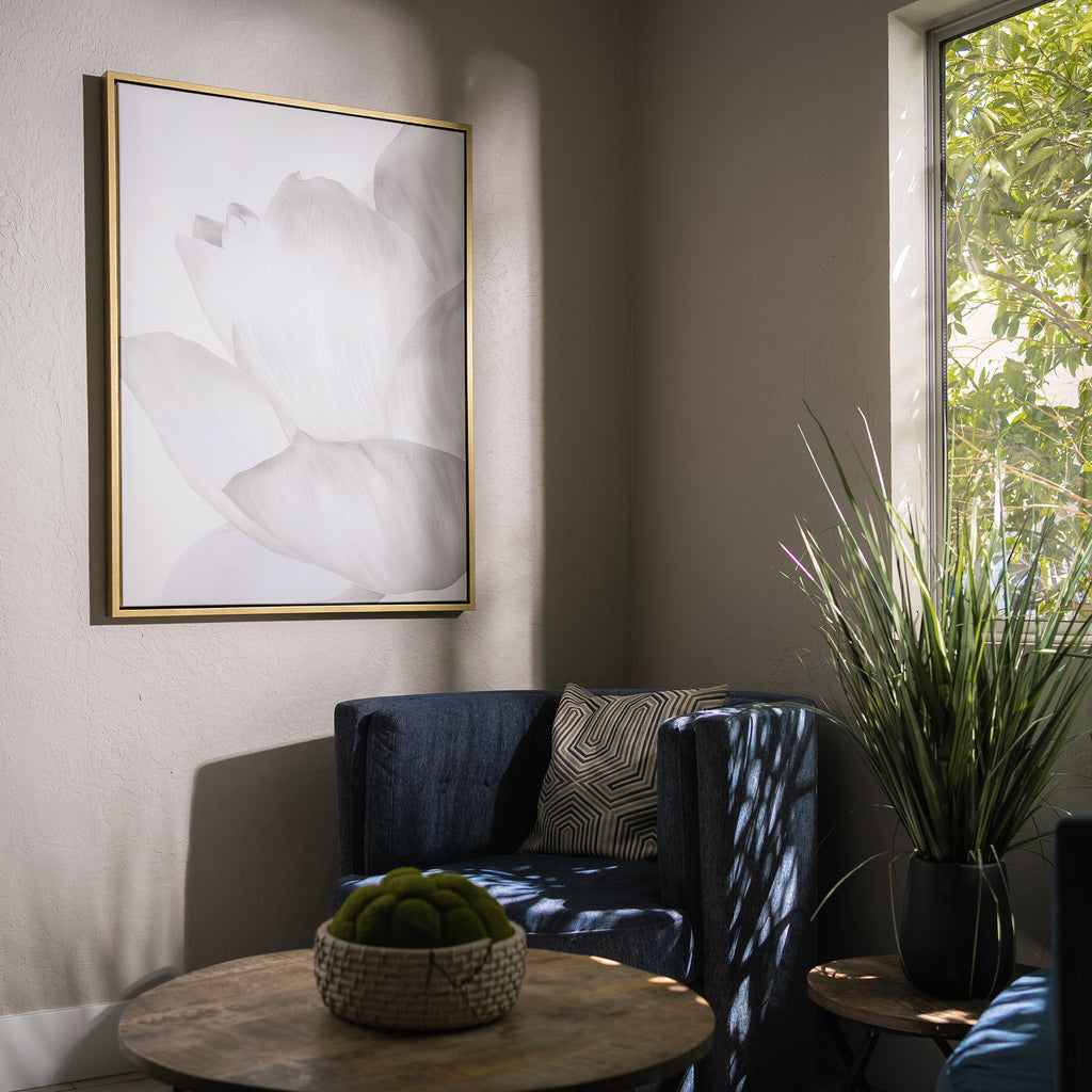 A cozy corner with a dark blue chair, patterned pillow, small round table topped with green moss balls, tall potted plant, and sunlight streaming in beneath the White Blossom Abstract Canvas Art.