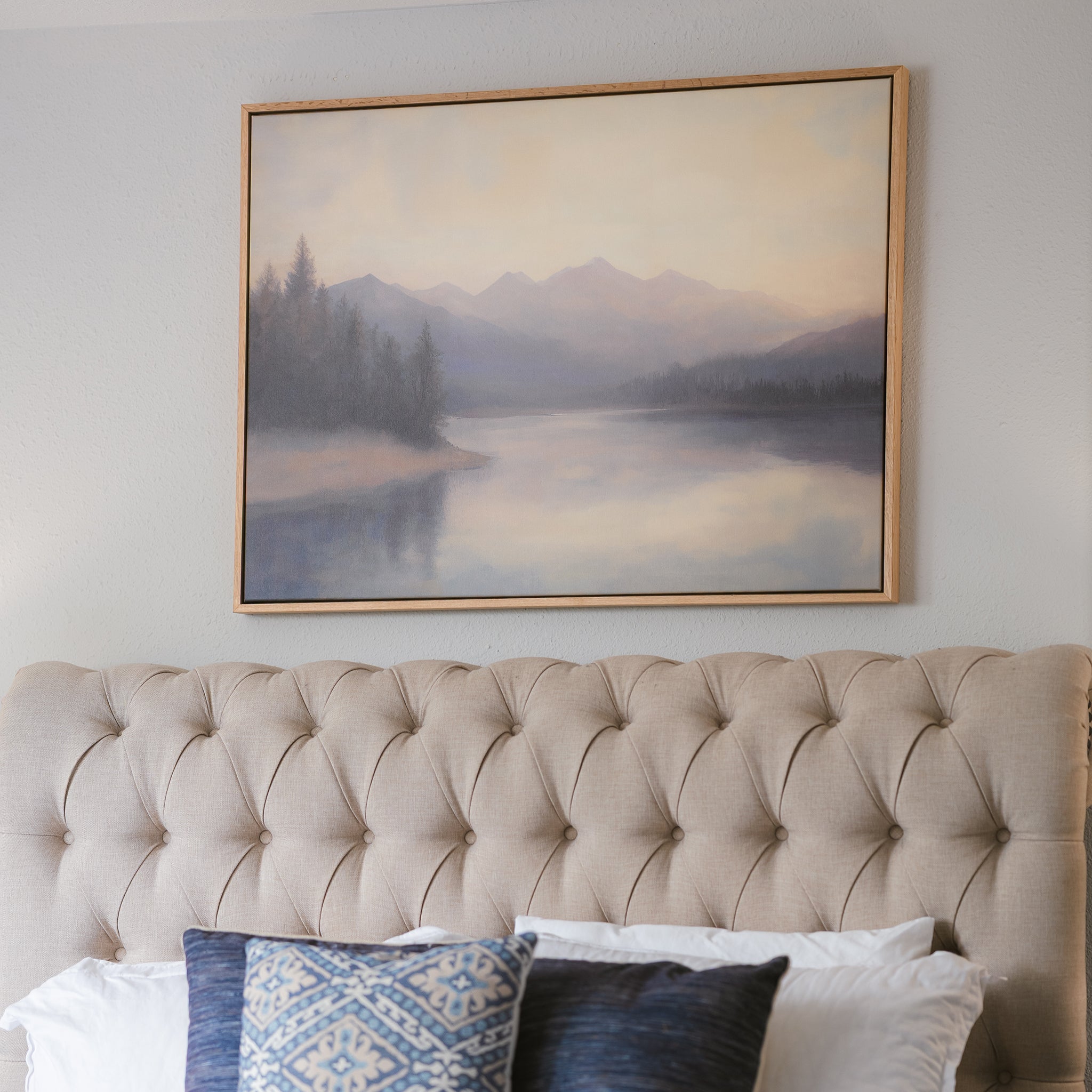 The Smoky Peaks Canvas Art, featuring misty mountains and a tranquil lake, hangs above a tufted beige headboard adorned with white and blue pillows.