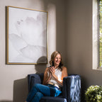 A woman sits in a blue armchair by a sunlit window, smiling and holding a mug. Behind her, White Blossom Abstract Canvas Art adds elegant minimalist wall decor, while a potted plant decorates the corner.
