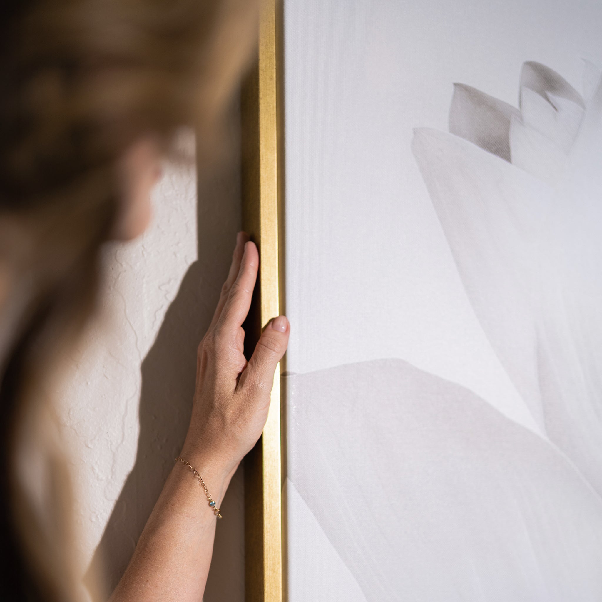 A hand gently touches the gold frame of the White Blossom Abstract Canvas Art, a minimalist floral print on a textured wall—only the hand, part of an arm, and blurred hair are visible, adding serenity to chic living room decor.