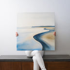 A person sits on a bench holding the Serene Coastline Canvas Art, a minimalist square painting featuring a tranquil shoreline with a winding path in muted blue and beige. Only the person's hands and legs are visible.