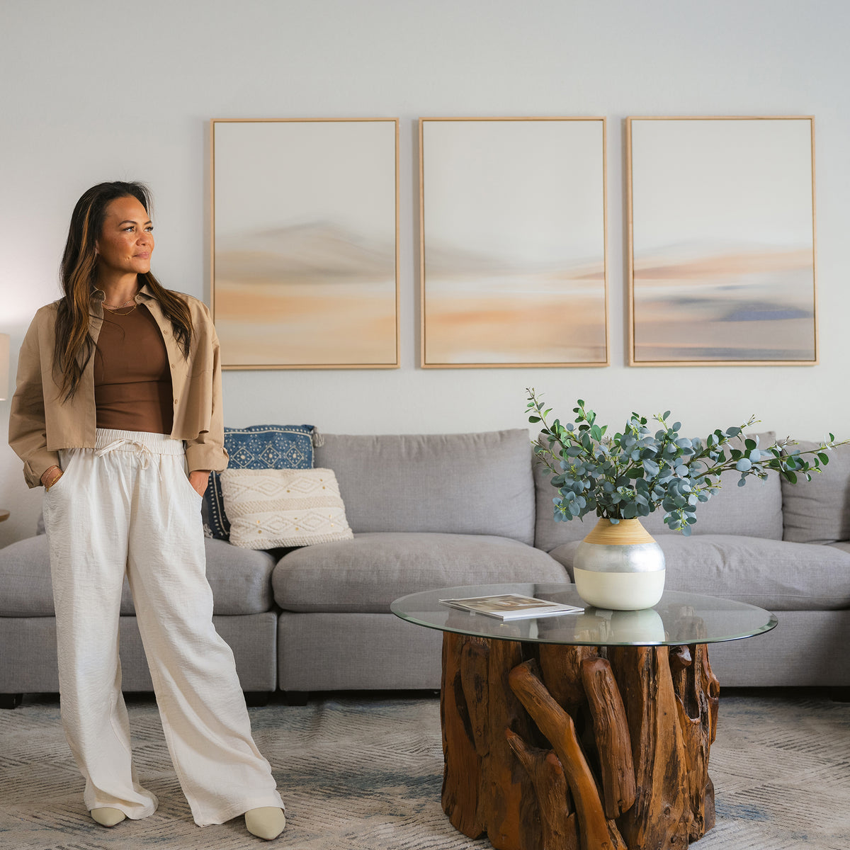 A woman stands in a chic living room, where the Desert Haze Set of 3 wall art hangs above a grey sofa. She wears a beige jacket, brown top, and white pants that echo the serene palette and style of the elegant decor.