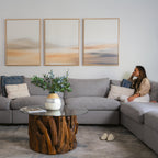 A woman sits on a gray sectional sofa in a modern living room with neutral decor, featuring the Desert Haze Set of 3 minimalist wall art prints, a glass coffee table with wood base, decorative pillows, and plants.