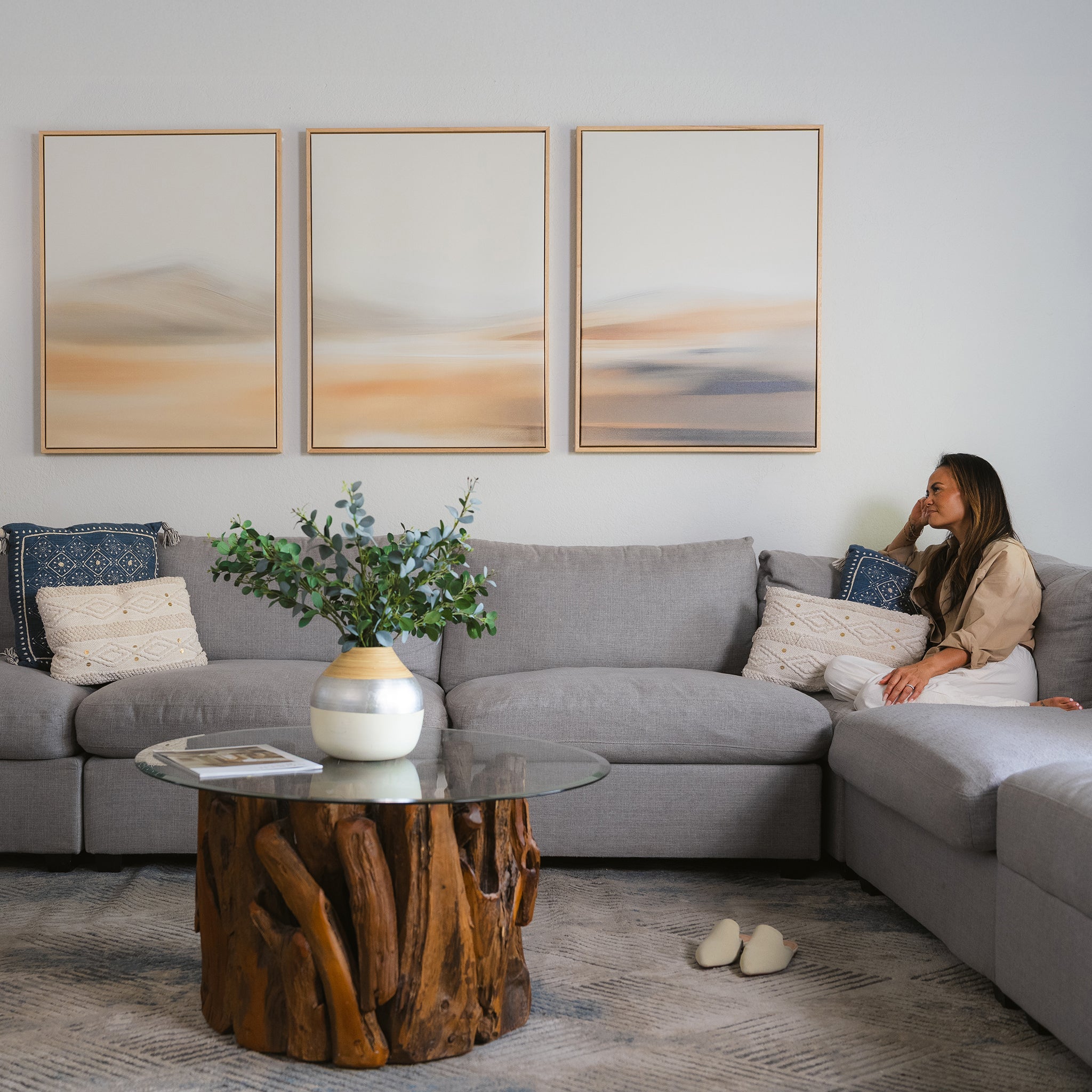 A woman sits on a gray sectional sofa in a modern living room with neutral decor, featuring the Desert Haze Set of 3 minimalist wall art prints, a glass coffee table with wood base, decorative pillows, and plants.