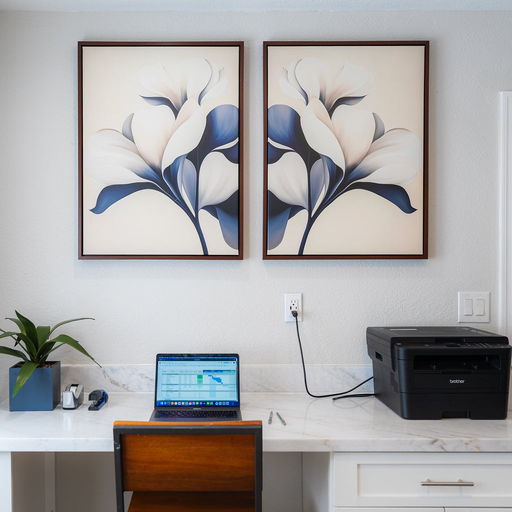 A modern home office setup with a laptop and supplies on a white marble desk features the Navy Magnolia Set of 2 as striking floral wall art above the workspace, adding elegant and modern decor to the scene.