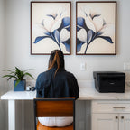 A person with long hair, in a dark shirt and white pants, sits at a white desk with a plant, pen holder, and printer. Above the desk, the Navy Magnolia Set of 2 wall art adds modern style to the scene.