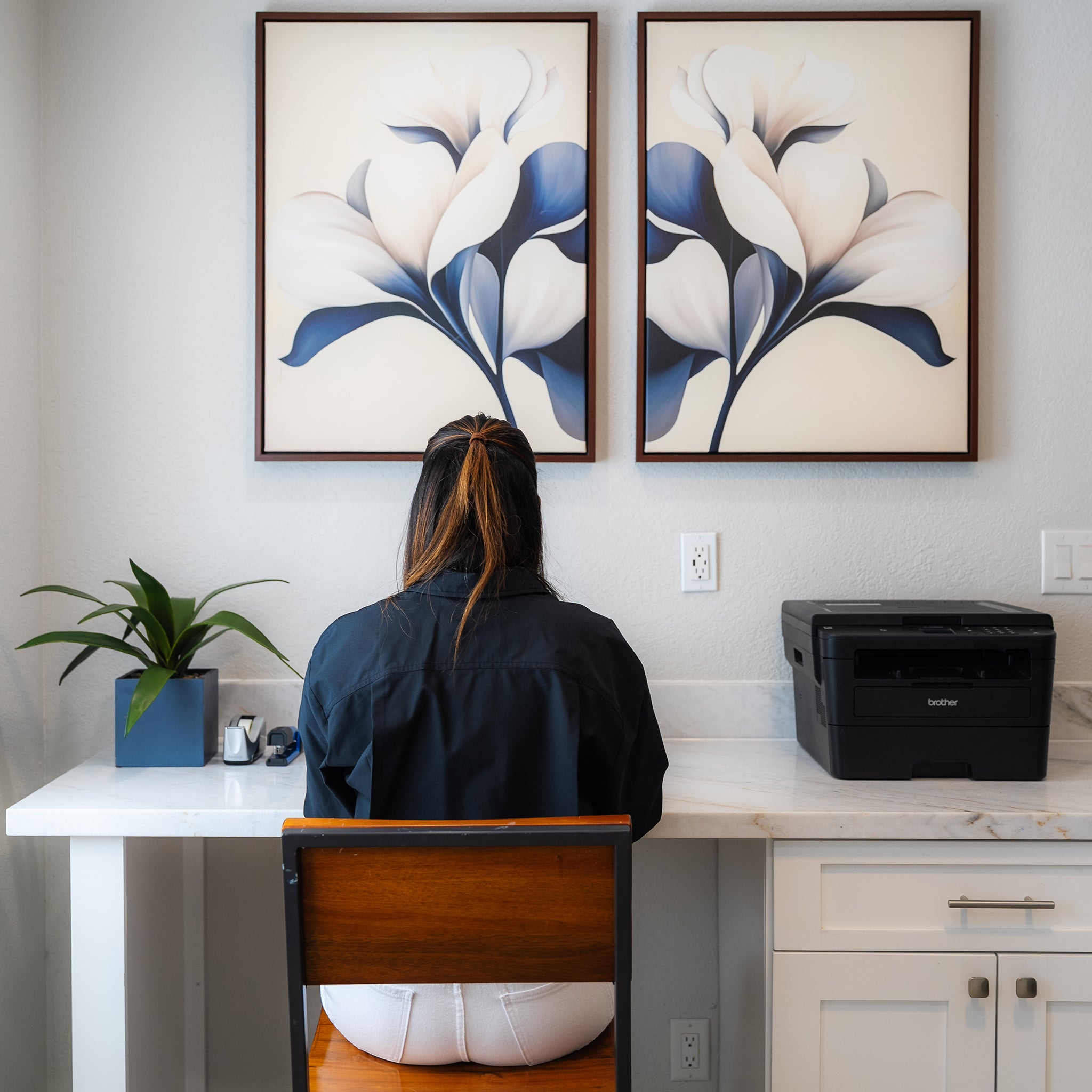 A person with long hair, in a dark shirt and white pants, sits at a white desk with a plant, pen holder, and printer. Above the desk, the Navy Magnolia Set of 2 wall art adds modern style to the scene.