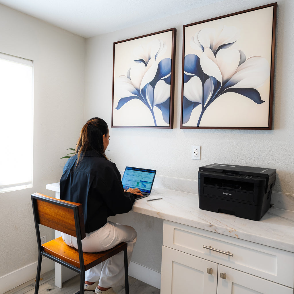 A woman works at a desk in a bright home office with white walls, a laptop, printer, plenty of natural light, and the Navy Magnolia Set of 2 as modern wall decor.