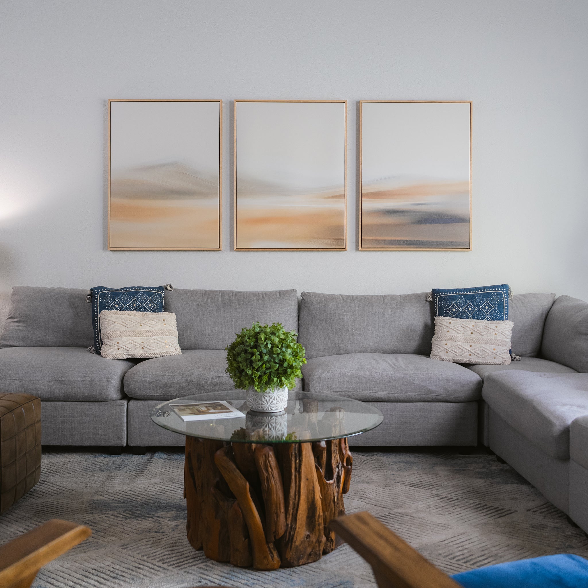 A modern living room features a gray sectional sofa, blue and cream pillows, a glass-topped coffee table with a wooden base, a potted plant, and the Desert Haze Set of 3 wall art above the sofa to enhance the elegant decor.