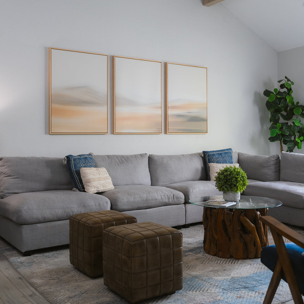 A modern living room features a large gray sectional, two brown ottomans, a round glass coffee table with a wood base, a potted plant, and the Desert Haze Set of 3 minimalist wall art above the sofa for stylish decor.