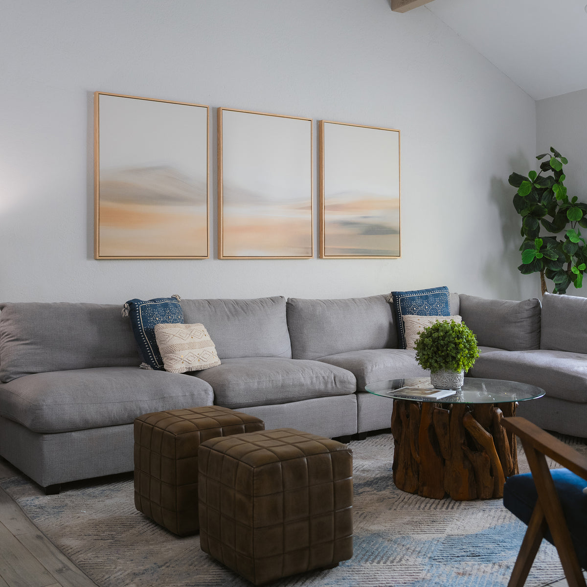 A modern living room features a large gray sectional, two brown ottomans, a round glass coffee table with a wood base, a potted plant, and the Desert Haze Set of 3 minimalist wall art above the sofa for stylish decor.